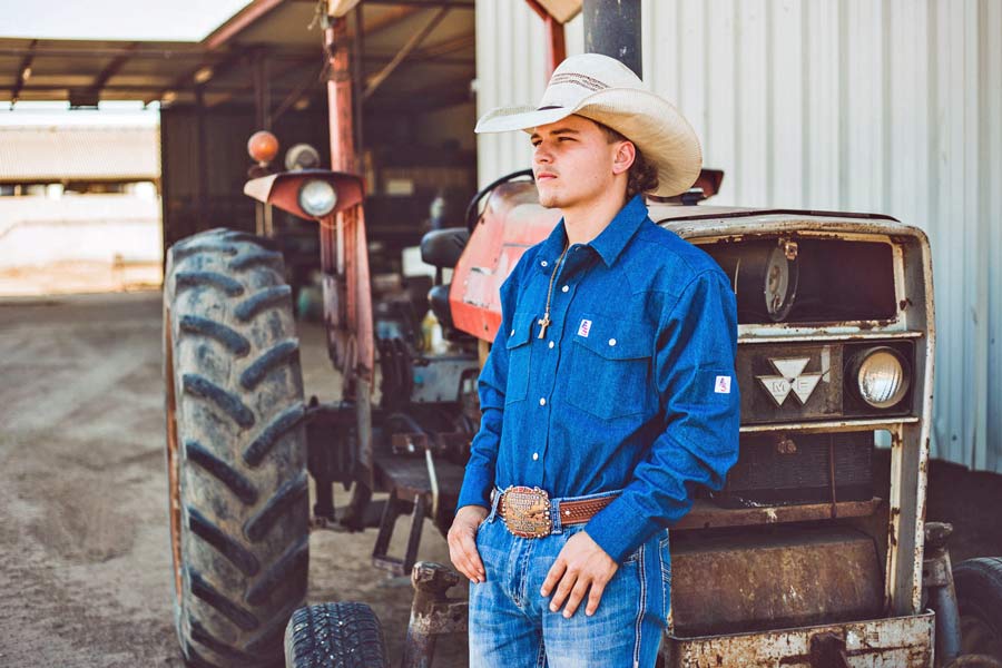 rodeo competitor thumbs in jean pockets with sponsored denim company branding photo shoot midsouth productions against tractor by cow barn with agriculture equipment near memphis tennessee