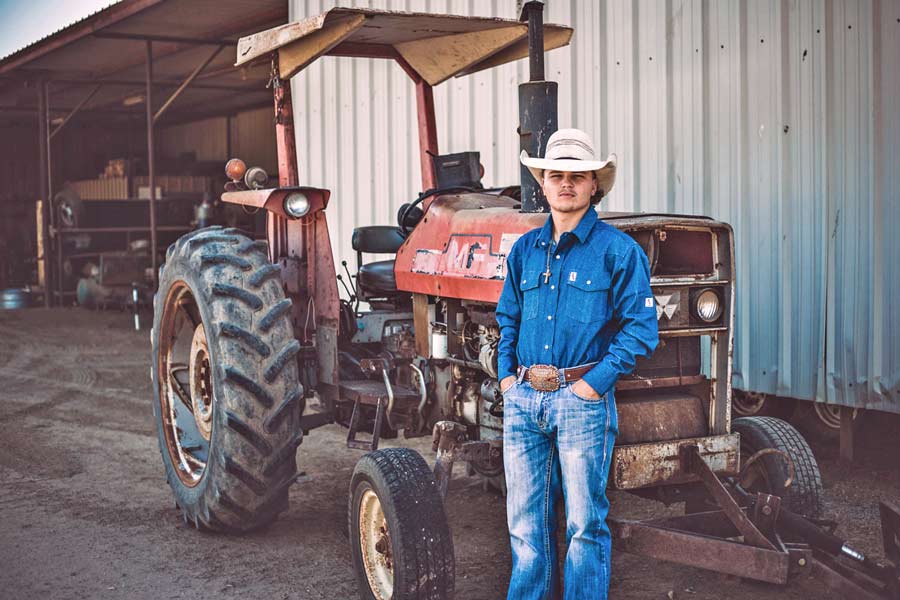 wide shot of bull rider leaning up against old red tractor in front of Midsouth cattle barn during creative photography session with prefocus for western website strategy