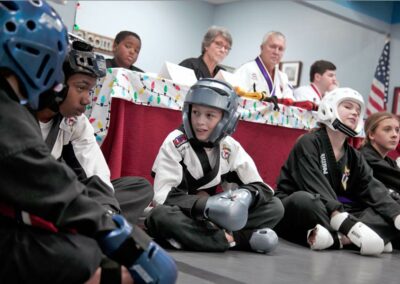 martial arts students seated in front of program owners and judges of taekwondo competition awards video services near memphis tn jordan trask of prefocus kids in head gear and sparring gloves